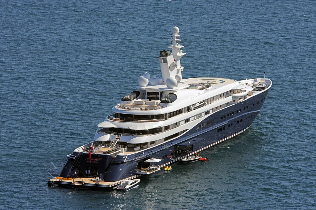 Al Mirqab at anchor seen from above and astern, navy hull, white superstructure, helipad on the foredeck and tenders at the stern platform