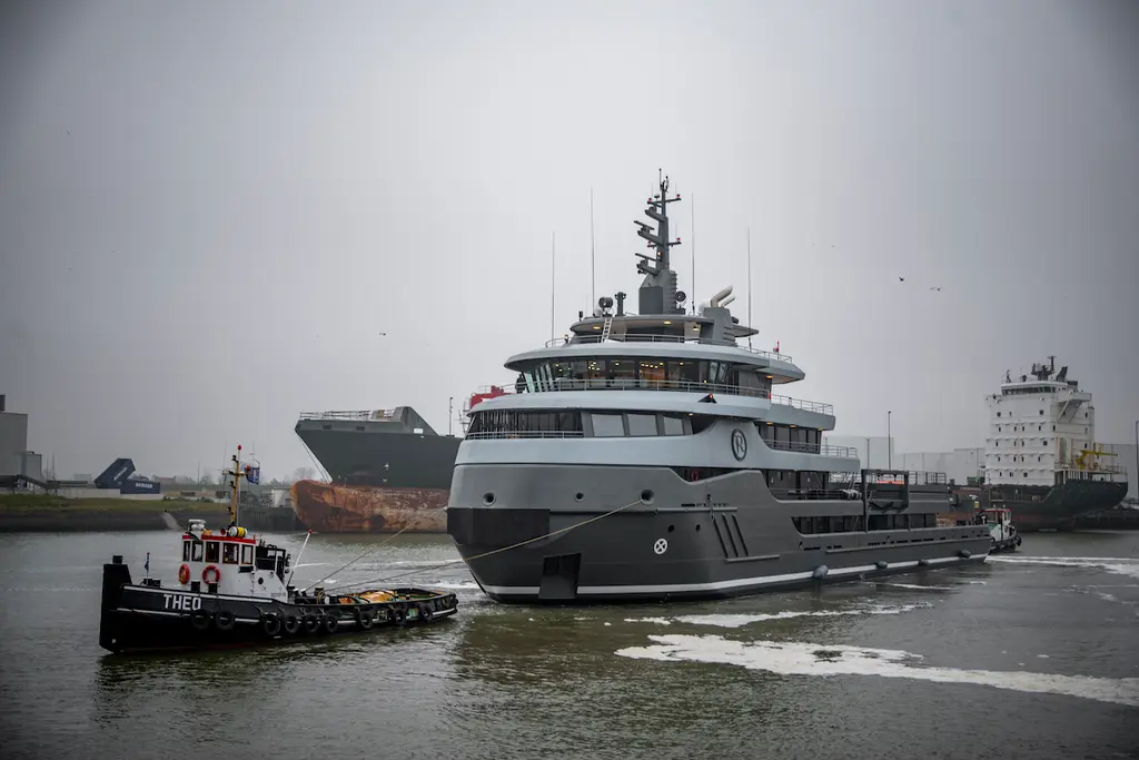 Ragnar in a grey industrial harbour with a small black harbour tug alongside and cargo ships behind