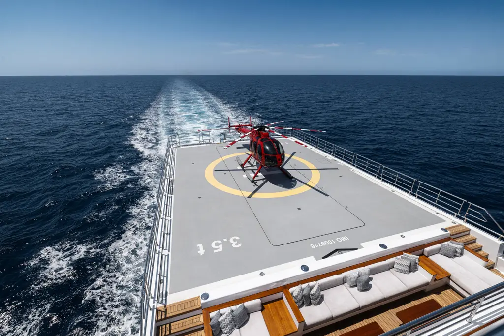 The aft helideck with a red helicopter on the yellow landing circle, the yacht's wake behind and a lounge with sofas at the forward edge of the deck