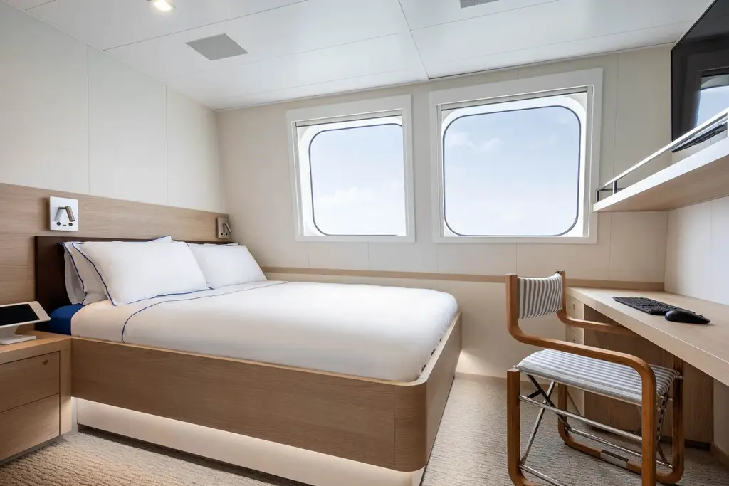 A cabin with a double bed, light oak panelling, two square windows and a desk with a chair