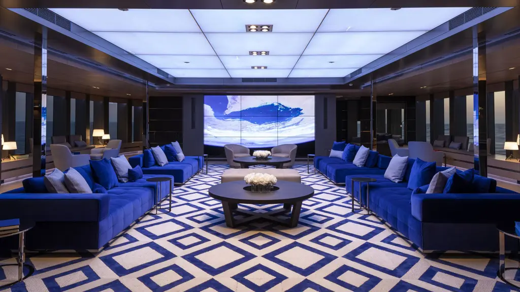 The main salon, a long lounge with blue sofas around a geometric blue and white carpet, under a backlit ceiling, with a large screen at the far end