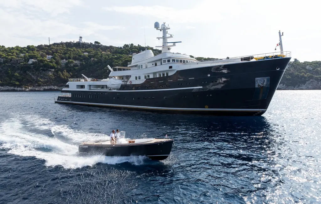 A dark-hulled tender speeding past the bow of Legend near a wooded coastline