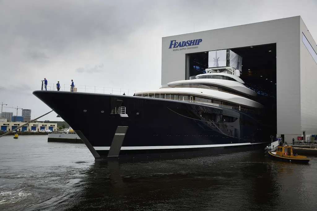 The bow of Breakthrough emerging from the Feadship build hall, with yard workers on the foredeck and a small yellow workboat alongside